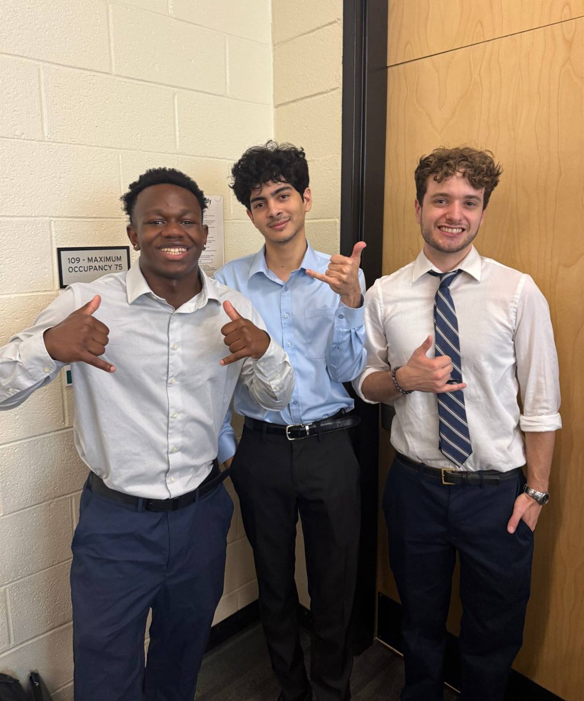 Three students in dress shirts stand together in a hallway, smiling and giving shaka signs