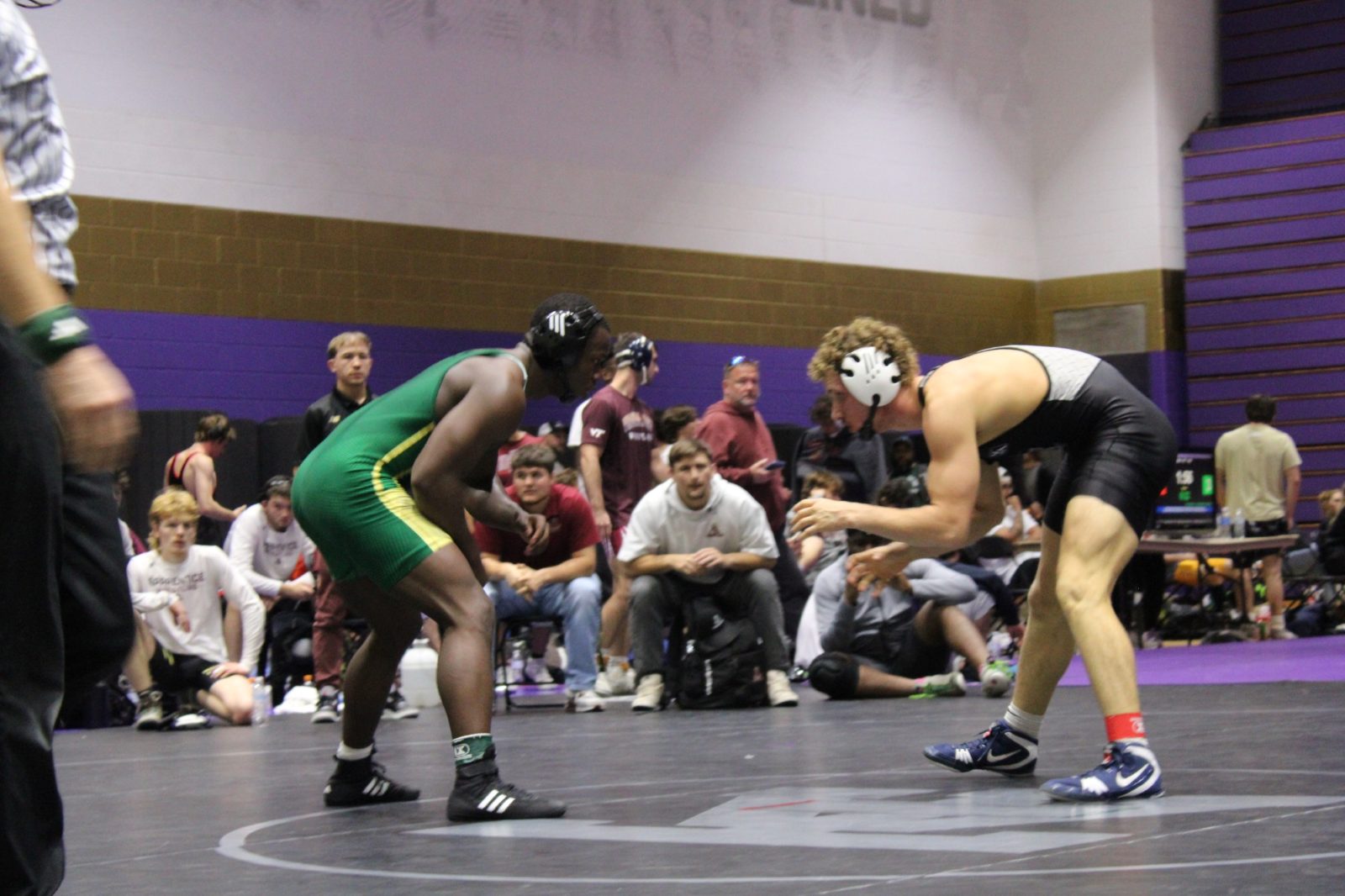 Two wrestlers square up on a black mat, one in a green and gold singlet and one in black, with teams watching from the edge of the mat